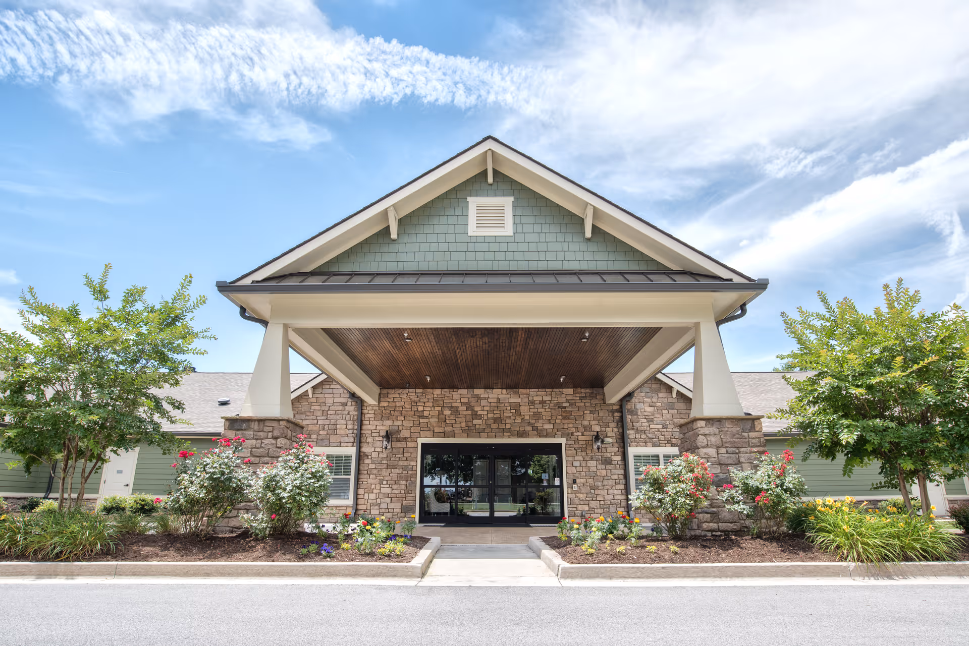 Front exterior view of a senior living facility with a covered entrance supported by stone pillars, surrounded by landscaped flower beds and small trees under a partly cloudy sky.