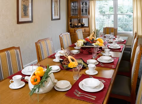 A long dining table set with white plates, cups, glasses, and silverware on red placemats. The table is decorated with fruit arrangements and a ceramic basket with bell peppers and greenery. The room has striped upholstered chairs, framed artwork on the wall, a wooden china cabinet, and large windows letting in natural light.