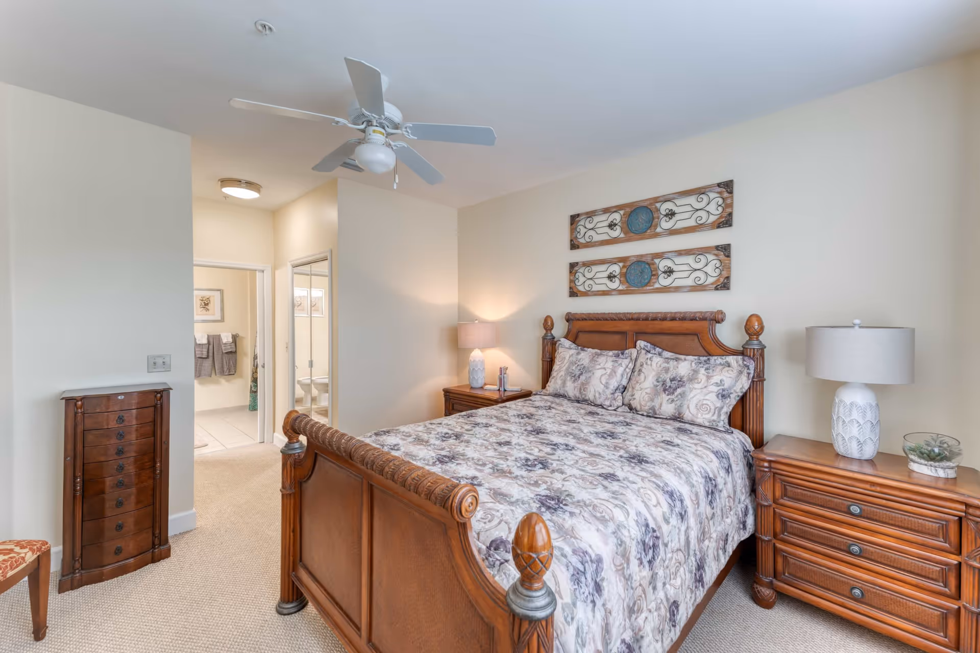 A bedroom with a wooden bed frame and floral patterned bedding. There are two matching wooden nightstands on either side of the bed, each with a lamp. Above the bed, two decorative wooden wall hangings are displayed. The room has beige walls and carpeted floor. In the background, there is a doorway leading to a bathroom with visible towels and a toilet.