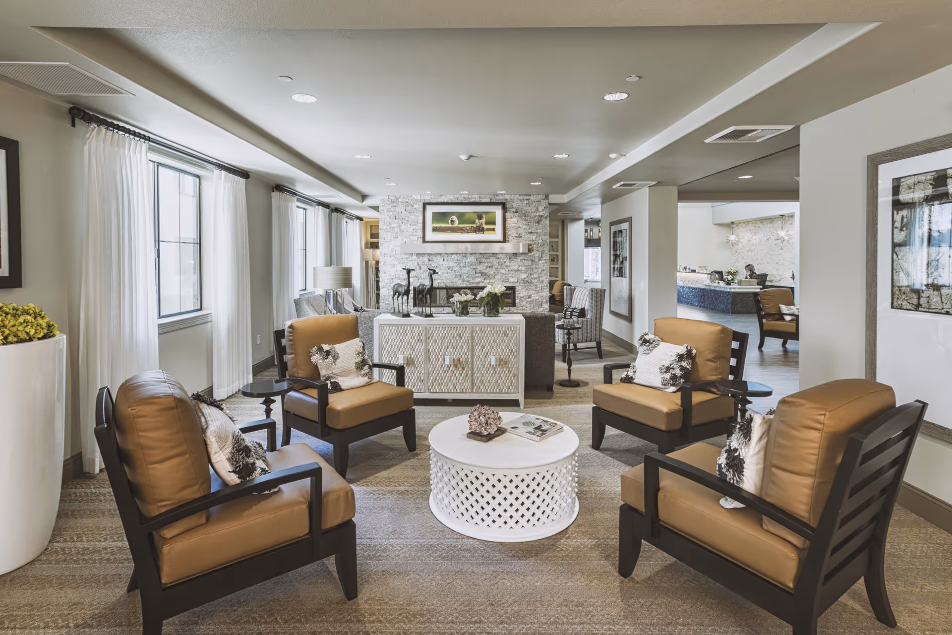 A cozy living room area with four brown cushioned armchairs arranged around a white round coffee table. The room features large windows with white curtains, a stone fireplace with a framed picture above it, and decorative items on a white cabinet behind the chairs. The space is well-lit with recessed ceiling lights and has neutral-toned walls and carpet.