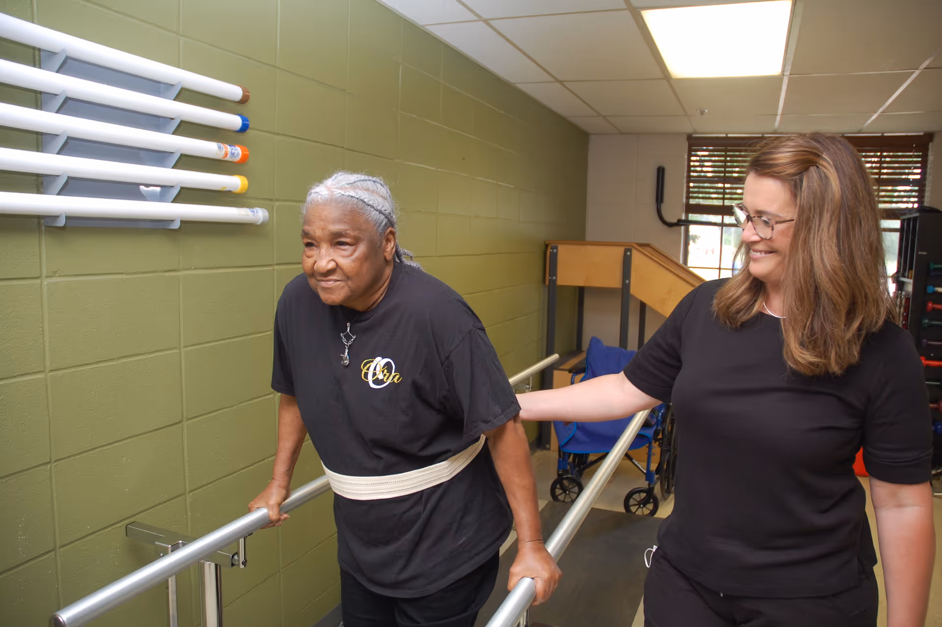 An elderly woman with gray braided hair is using parallel bars for physical therapy while a female therapist in black assists and supports her. The setting appears to be a rehabilitation or therapy room with a green wall and exercise equipment in the background.