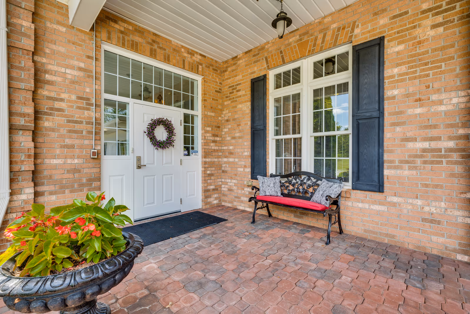 Covered brick front porch with a white entry door decorated with a wreath, a bench with a red cushion, and a potted plant.