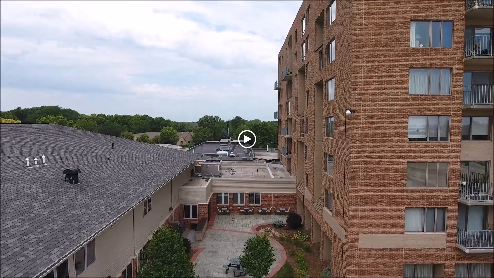 Aerial view of a senior living facility showing a multi-story brick building on the right and a lower building with a gray roof on the left. There is a courtyard area with trees, benches, and a walking path between the buildings. In the background, there are trees and houses under a partly cloudy sky.