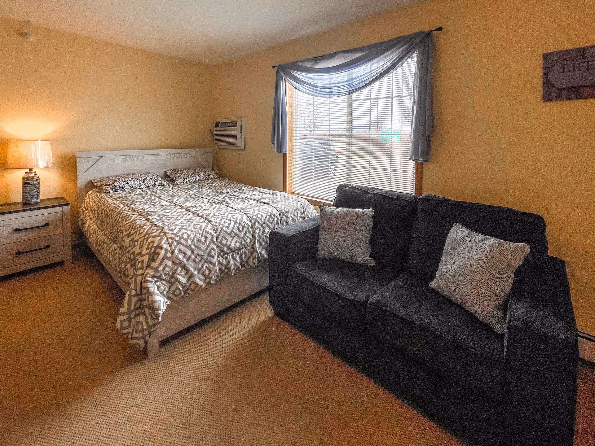 A cozy bedroom with a double bed covered in a patterned comforter, a nightstand with a lamp, a window with blinds and a sheer blue curtain, and a dark sofa with two patterned pillows.