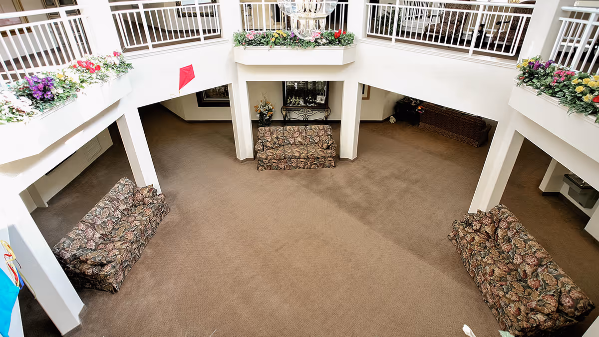 Spacious two-story atrium lounge with patterned sofas, carpeted floor, and flower boxes along the upper railing.