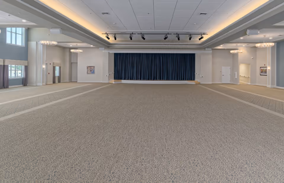 A large, empty event room with patterned carpet flooring, a stage with closed dark blue curtains, multiple chandeliers hanging from the ceiling, and large windows on the left side allowing natural light to enter.