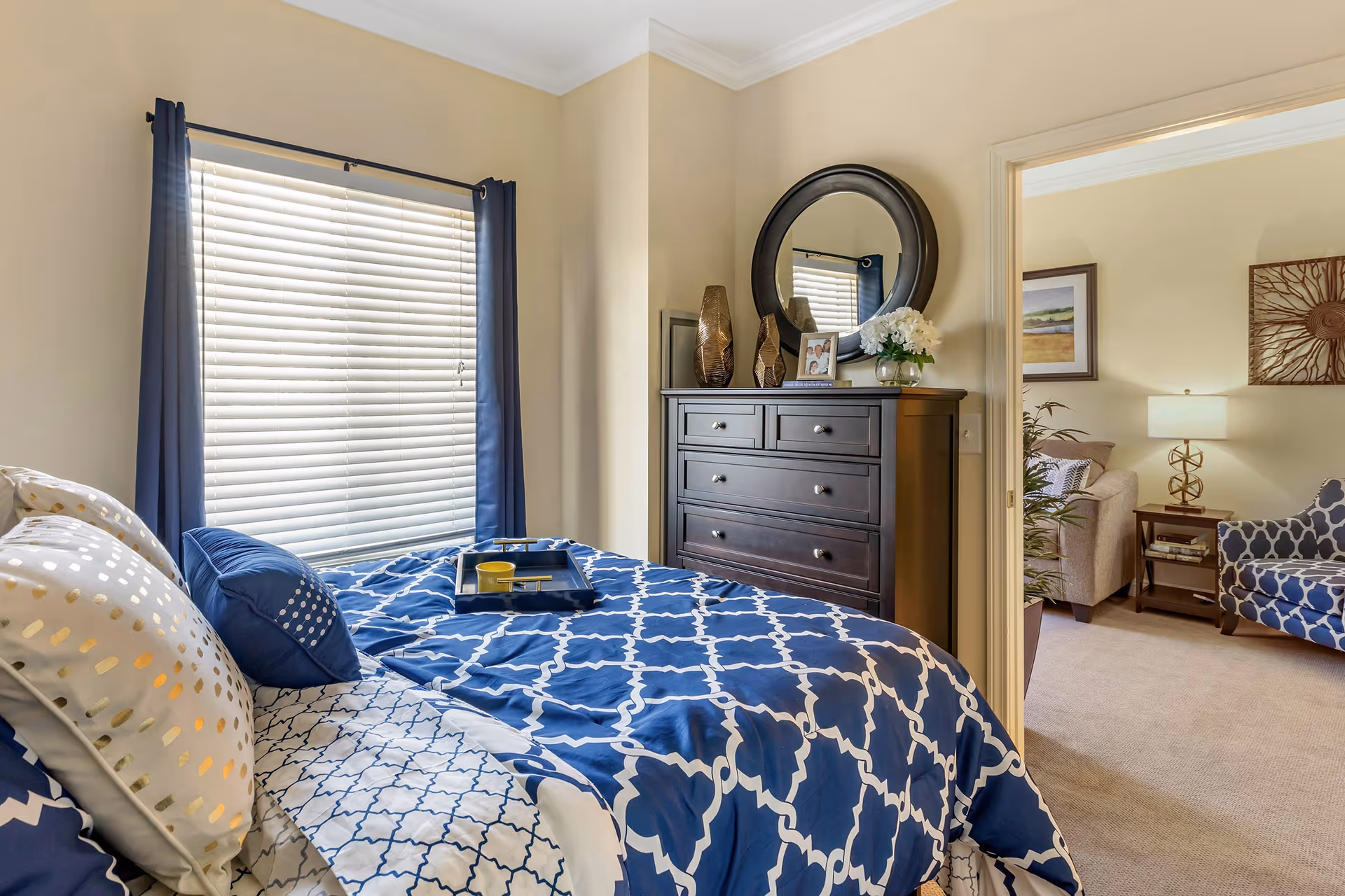 A bedroom with a bed covered in blue and white patterned bedding, a window with closed blinds and blue curtains, and a dark wooden dresser with a round mirror above it. The dresser has decorative vases, a framed photo, and a small flower arrangement. An open doorway reveals a living room area with a beige sofa, a blue patterned armchair, a side table with a lamp, and wall decorations.
