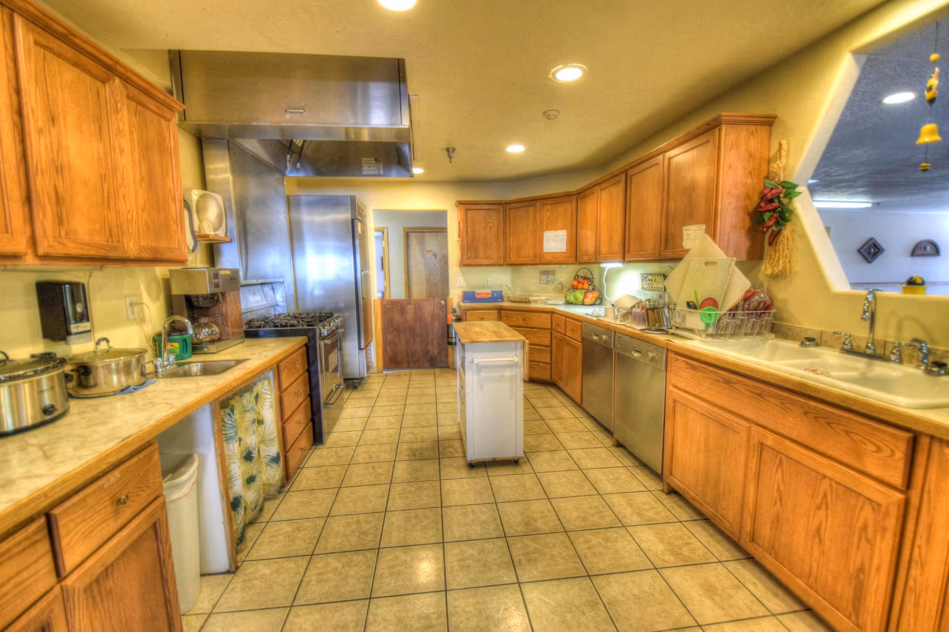 A large kitchen with wooden cabinets, tiled floor, and stainless steel appliances including a stove, refrigerator, and dishwasher. There is a small island in the center and a double sink under a window opening to an adjacent room. Various kitchen items and utensils are visible on the counters.