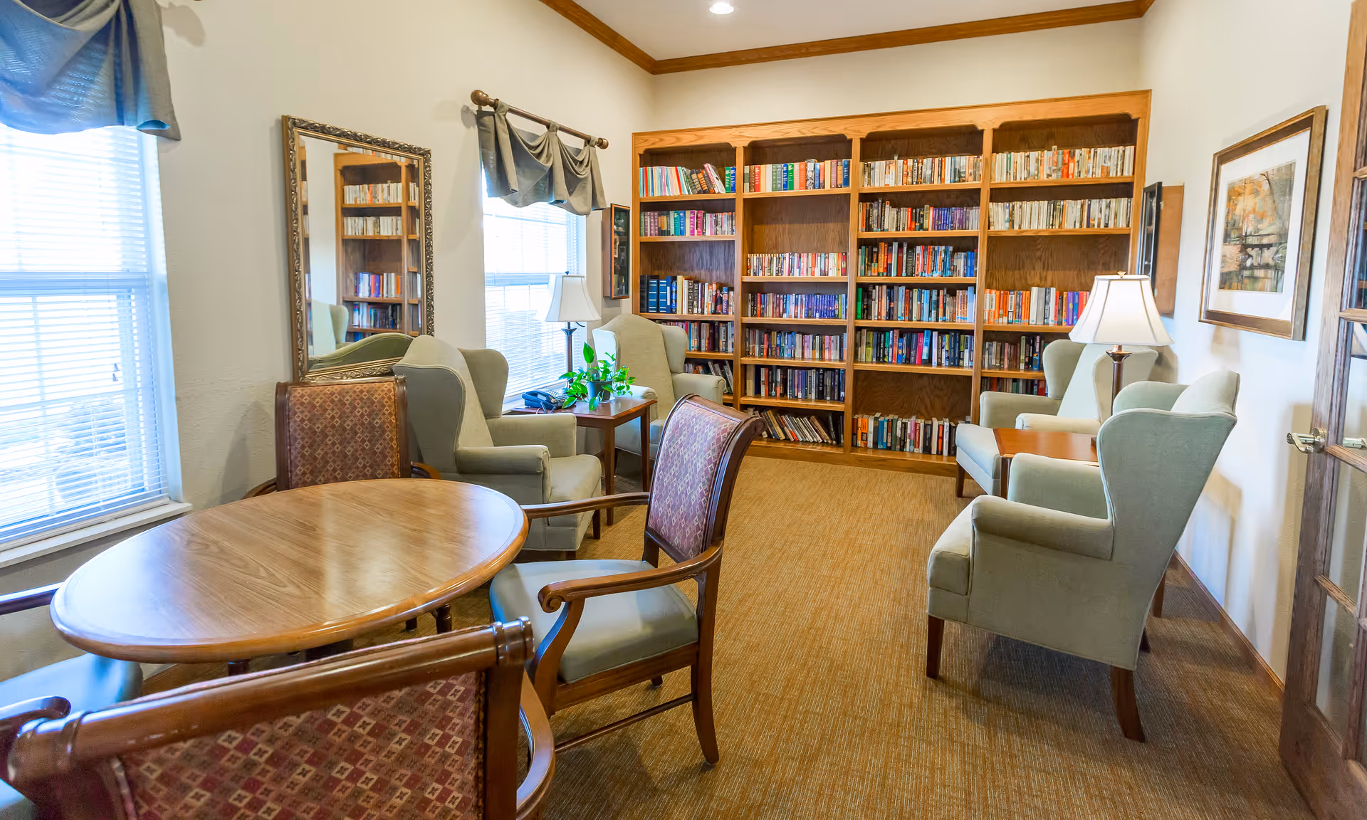 A cozy reading room with wooden bookshelves filled with books along the back wall. There are four green upholstered armchairs arranged around two wooden side tables with lamps. A round wooden table with four patterned chairs is in the foreground. Two windows with blinds and valances let in natural light. A large mirror hangs on the left wall, and framed artwork is on the right wall.