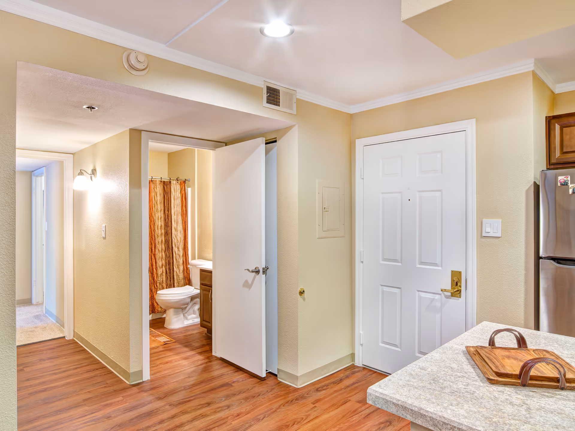 Interior view of a senior living facility showing a hallway with a bathroom door open, revealing a toilet and shower curtain. The hallway has wooden flooring and beige walls with a white ceiling light. A white entrance door is visible next to a kitchen counter with a wooden tray on it, and a stainless steel refrigerator is partially visible on the right.