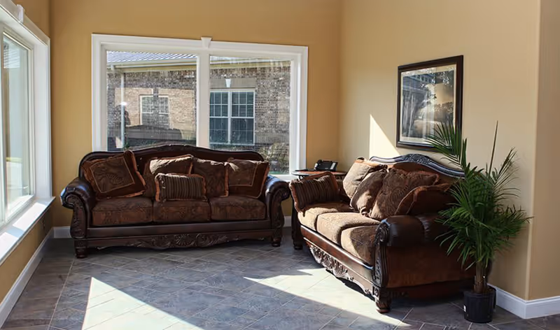 Two brown upholstered sofas arranged in a sunlit seating area with large windows, tile floor, framed art, and a potted plant.