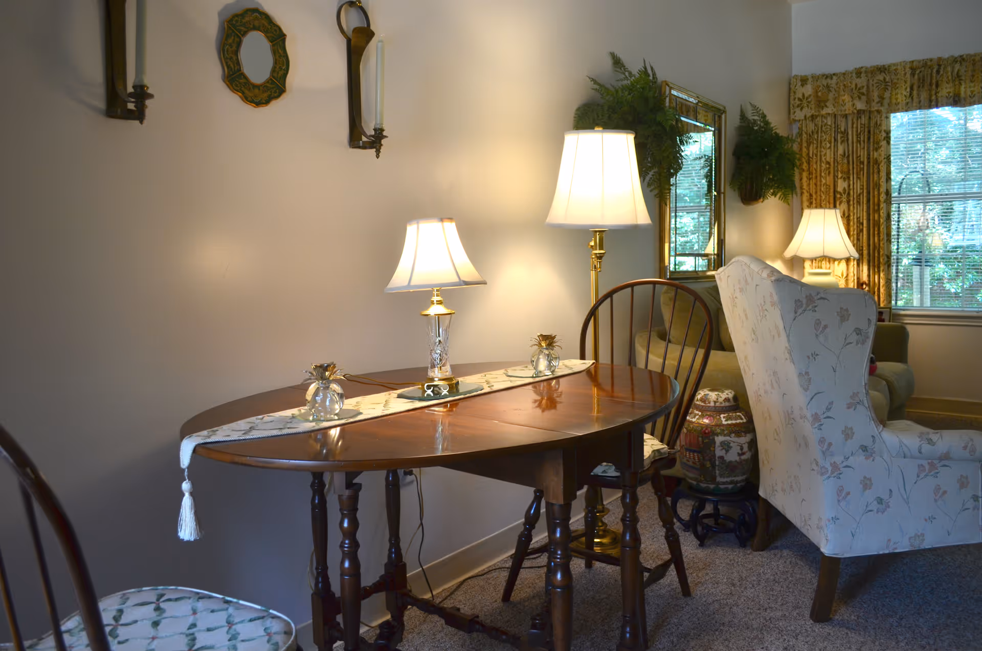 A cozy interior living space featuring a wooden table with a decorative runner and a small lamp on it. There are wooden chairs around the table, a floral upholstered armchair, a floor lamp, and a window with patterned curtains letting in natural light. The walls have decorative sconces and a small mirror with greenery above it.