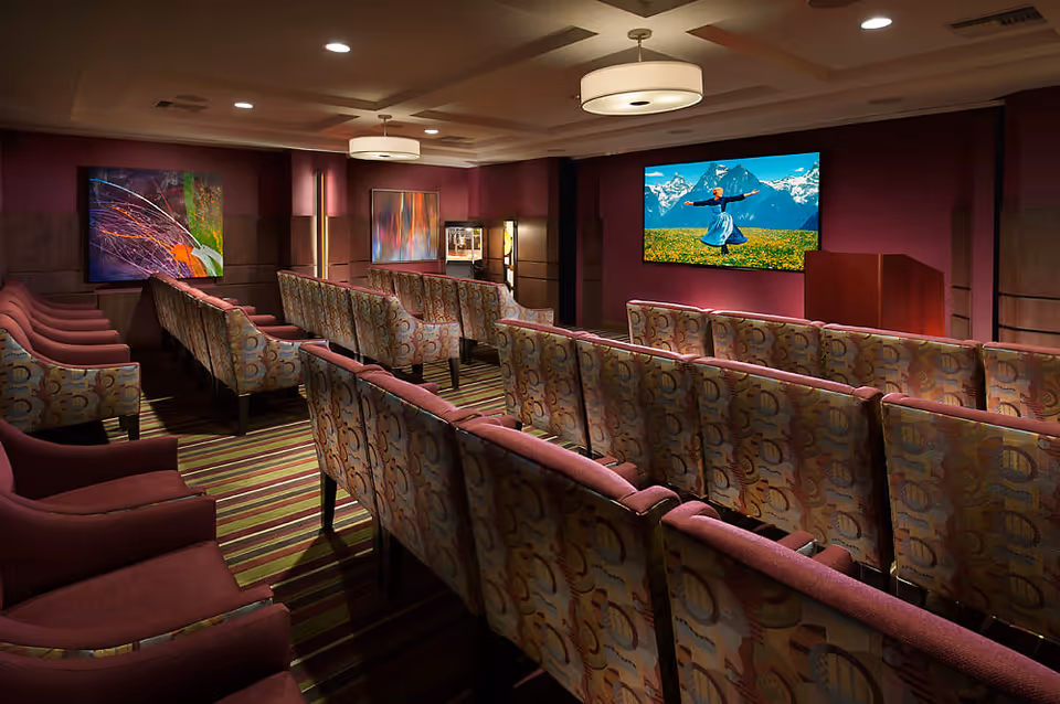 A cozy theater room with multiple rows of patterned and solid upholstered chairs facing a large screen displaying a scene from a movie with mountains and a person dancing in a field. The room has warm lighting, striped carpet, and colorful abstract paintings on the walls.