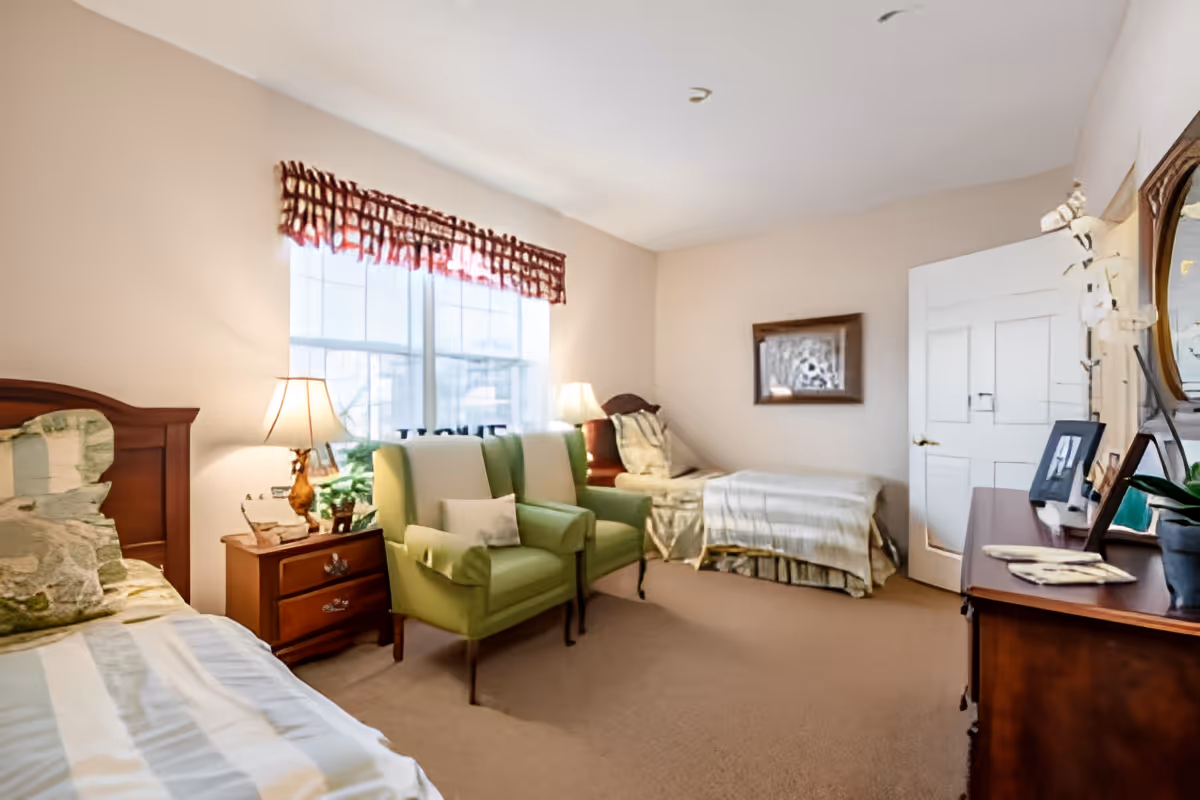 A cozy bedroom with two single beds, a wooden nightstand with a lamp between them, two green armchairs in front of a large window with a valance, a wooden dresser with a mirror and decorative items, and a closed white door.