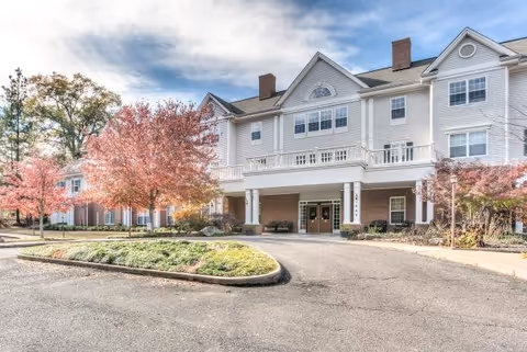 Exterior view of a large, multi-story assisted living facility with white siding and brick accents. The building has multiple windows, a covered entrance with double doors, and a circular driveway with landscaped greenery and trees with autumn foliage.