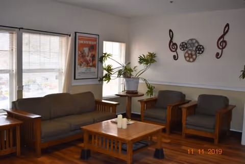 A cozy living room with wooden furniture including a sofa, two armchairs, a coffee table with candles, and side tables. There are large windows with blinds letting in natural light, a potted plant on a small round table, and wall decorations featuring musical notes and film reels. A framed poster is visible on the wall near the windows.
