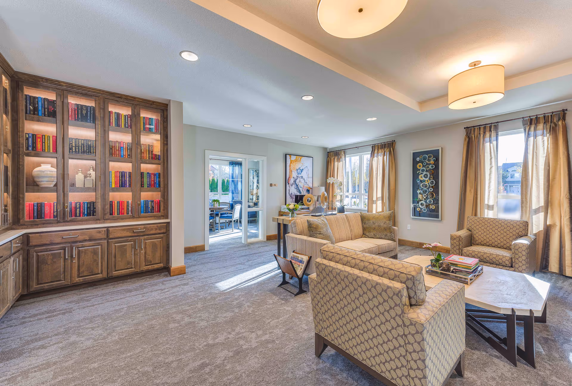 A bright and spacious living room with beige patterned armchairs and a matching sofa arranged around a wooden coffee table. The room features large windows with beige curtains allowing natural light to fill the space. A built-in wooden bookshelf filled with colorful books and decorative items is on the left wall. The walls are decorated with modern artwork, and the ceiling has recessed lighting and two round ceiling lights.