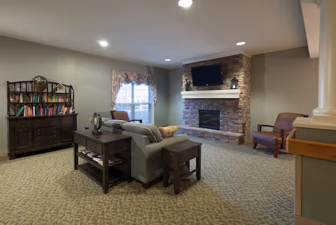 Cozy common room with a sofa facing a stone fireplace and wall-mounted TV, bookcase and additional chairs.