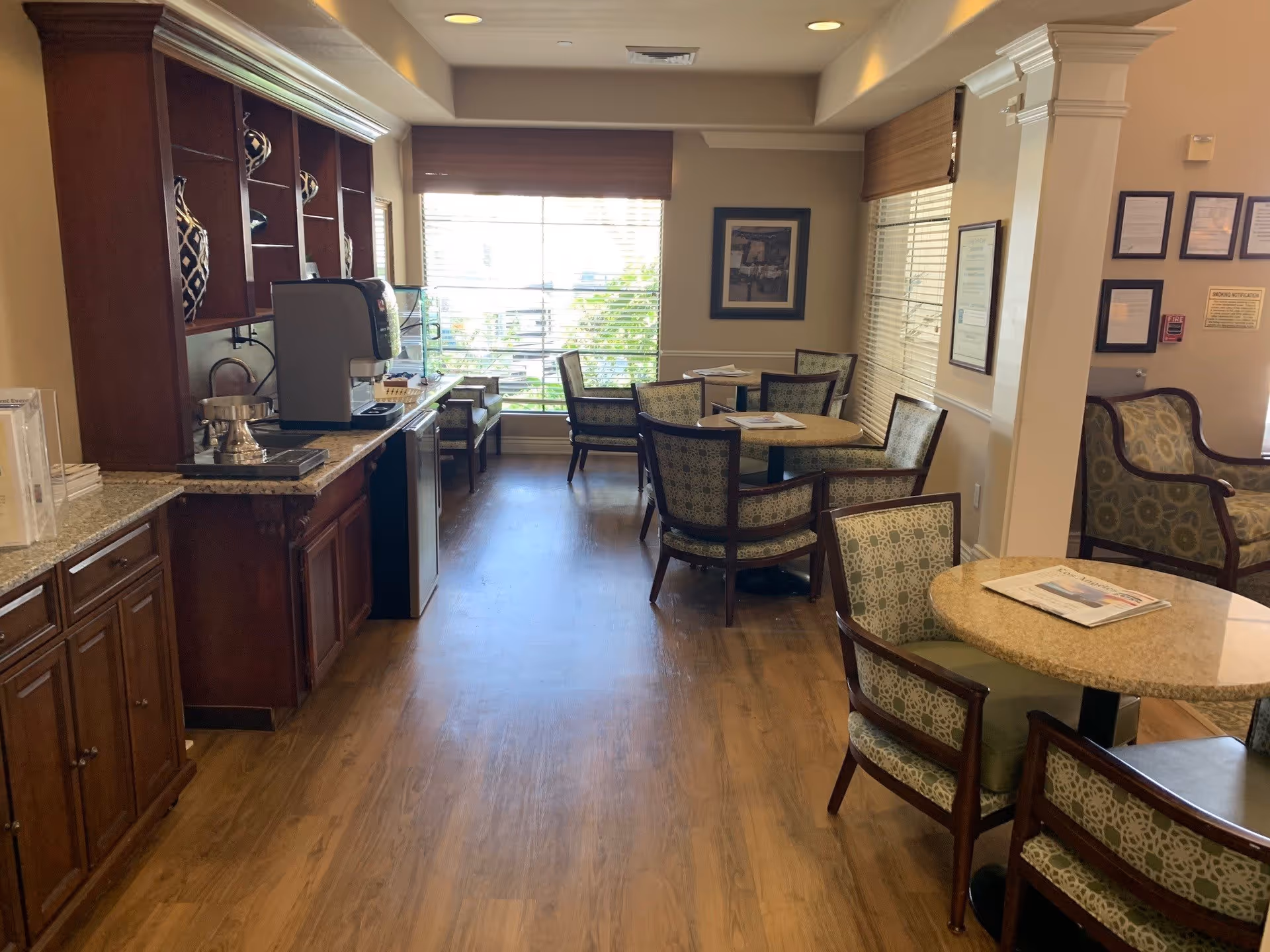 A cozy dining area in a senior living facility with wooden flooring, round tables with patterned chairs, a coffee machine on a granite countertop, and large windows with blinds letting in natural light.
