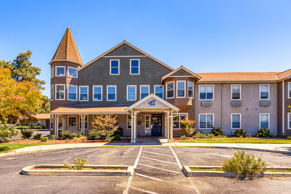 Front exterior view of a senior living facility building with a peaked roof and a small tower on the left side. The building has multiple windows and a covered entrance with a small porch. There are landscaped bushes and trees around the entrance and a parking area in front.