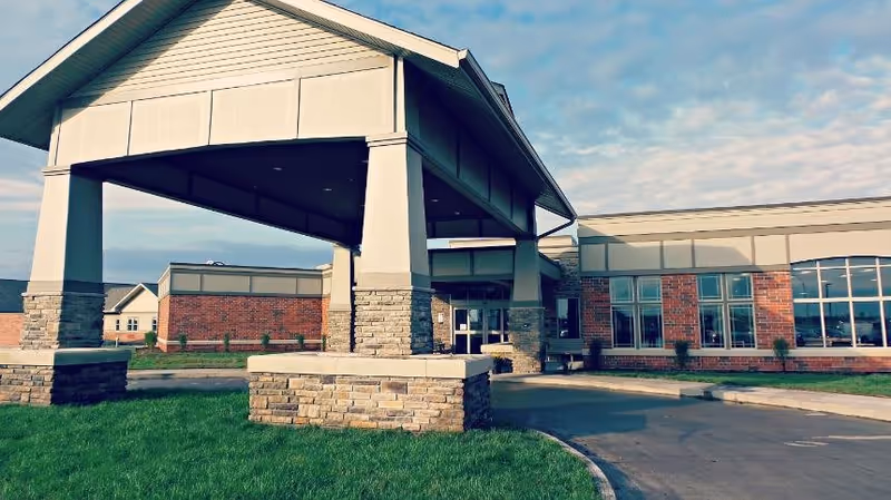 Exterior view of a senior living facility building with a covered entrance supported by large pillars made of stone and siding. The building has large windows and a brick facade, with a driveway curving in front of the entrance and green grass surrounding the area under a partly cloudy sky.