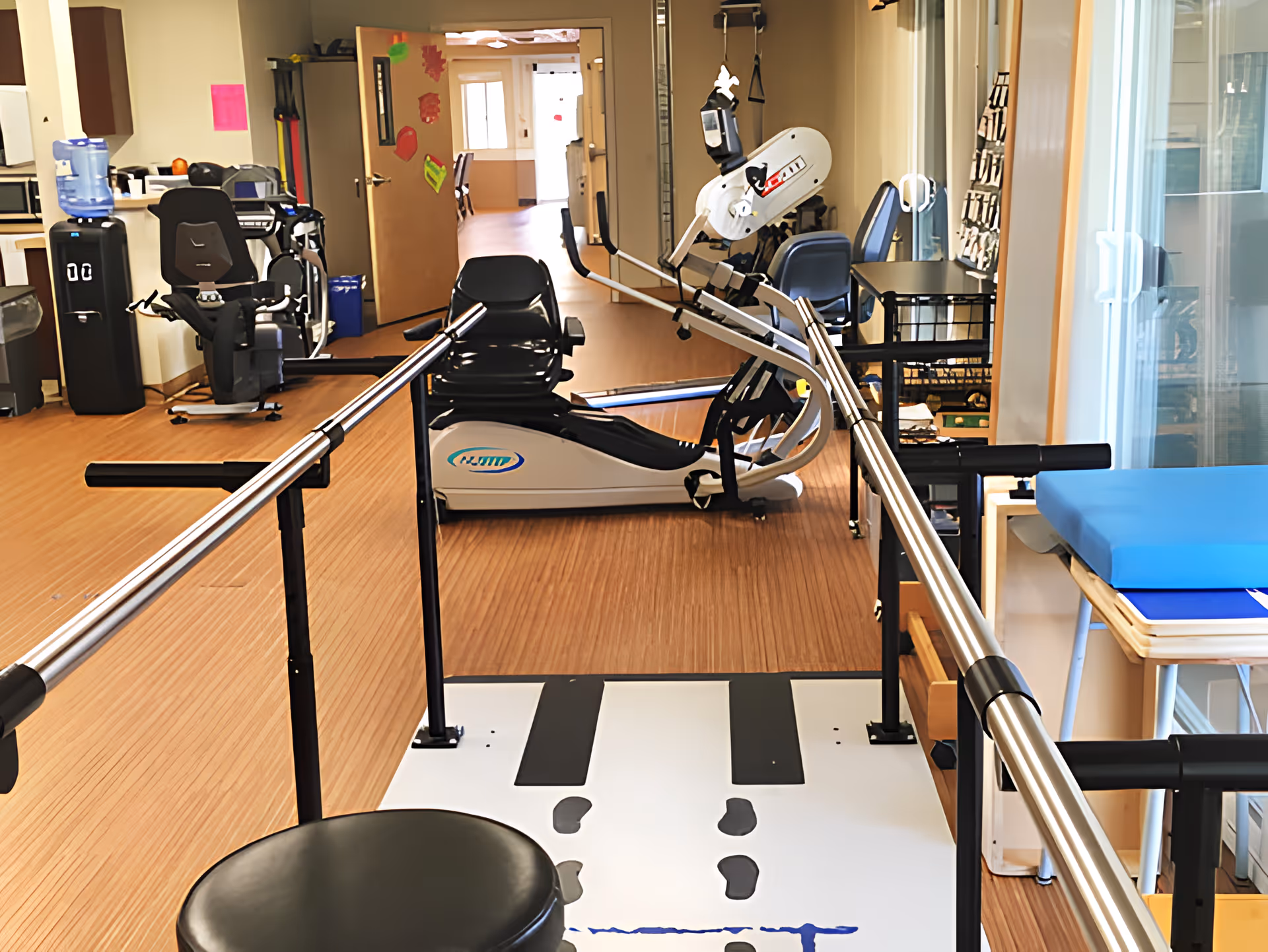 Physical therapy room with parallel bars for walking exercises, a recumbent exercise bike, a water cooler, chairs, and a treatment table with a blue cushion. The room has wood flooring and an open door leading to a hallway.