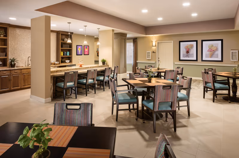 Well-lit assisted living dining room with multiple tables and chairs, a long counter with barstools, and framed artwork on the walls.