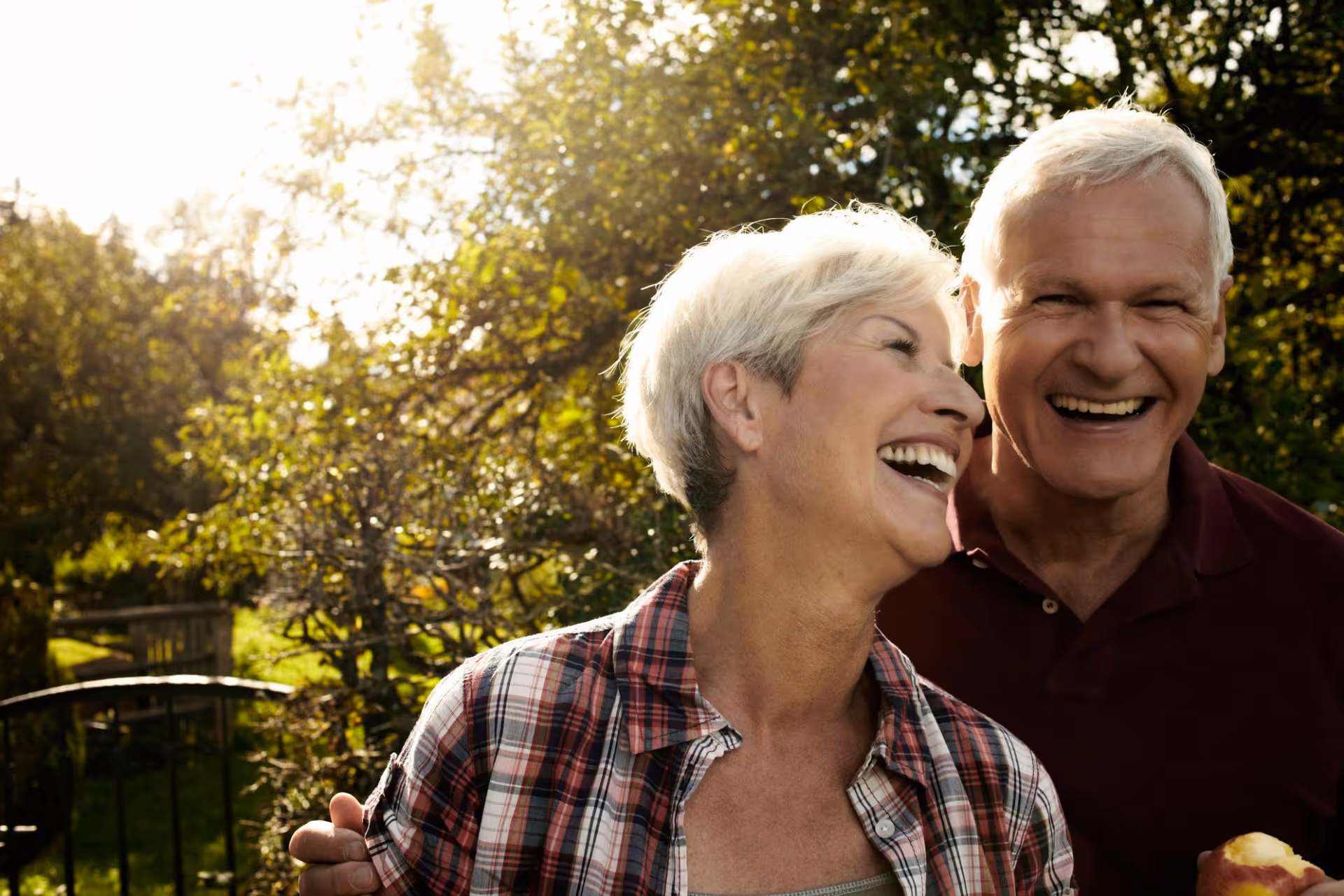 An older couple laughing together outdoors in a sunlit garden.