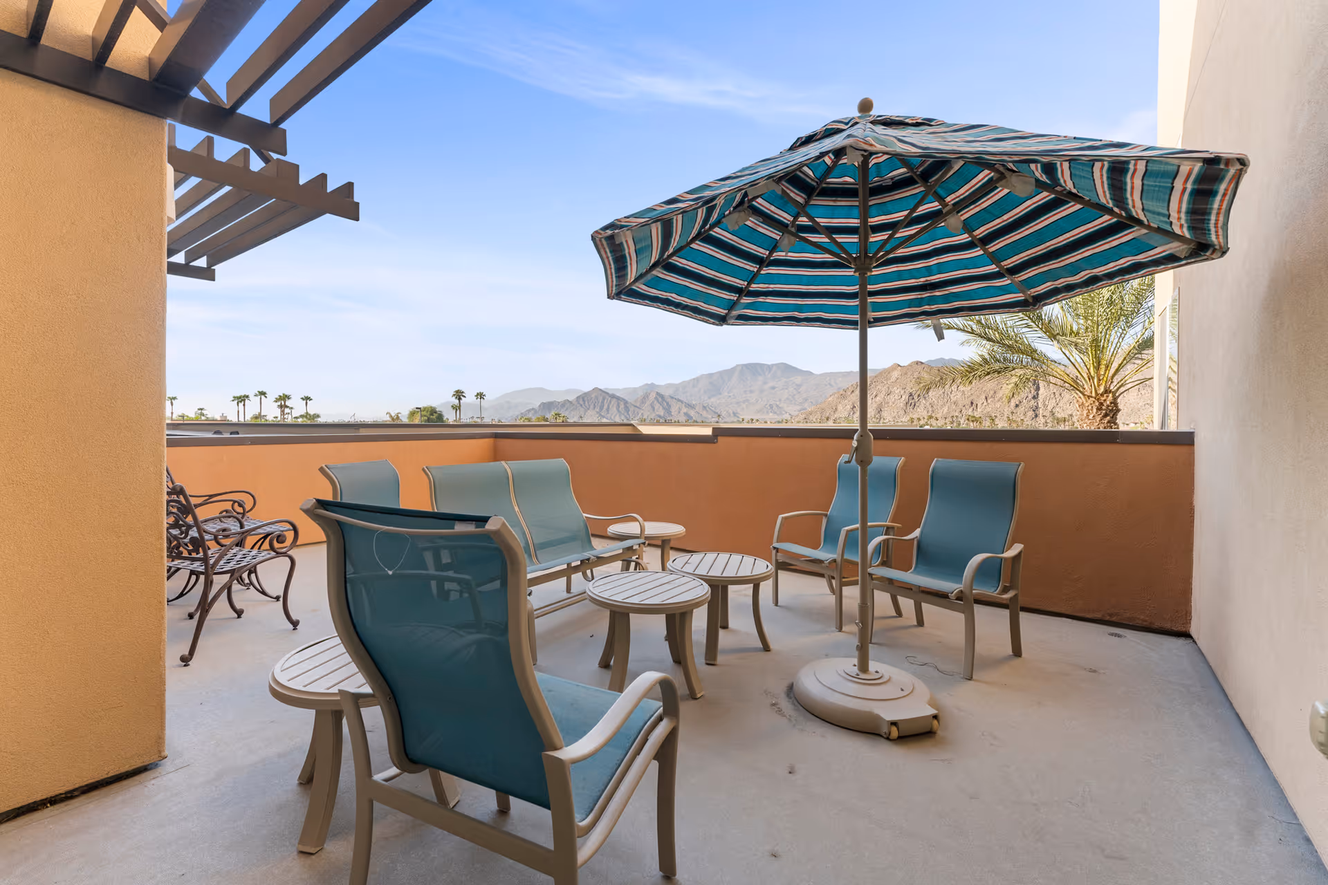 Outdoor patio area with several blue cushioned chairs arranged around small round tables, and a large striped umbrella providing shade. The patio is enclosed by a low wall with a view of distant mountains and palm trees under a clear blue sky.