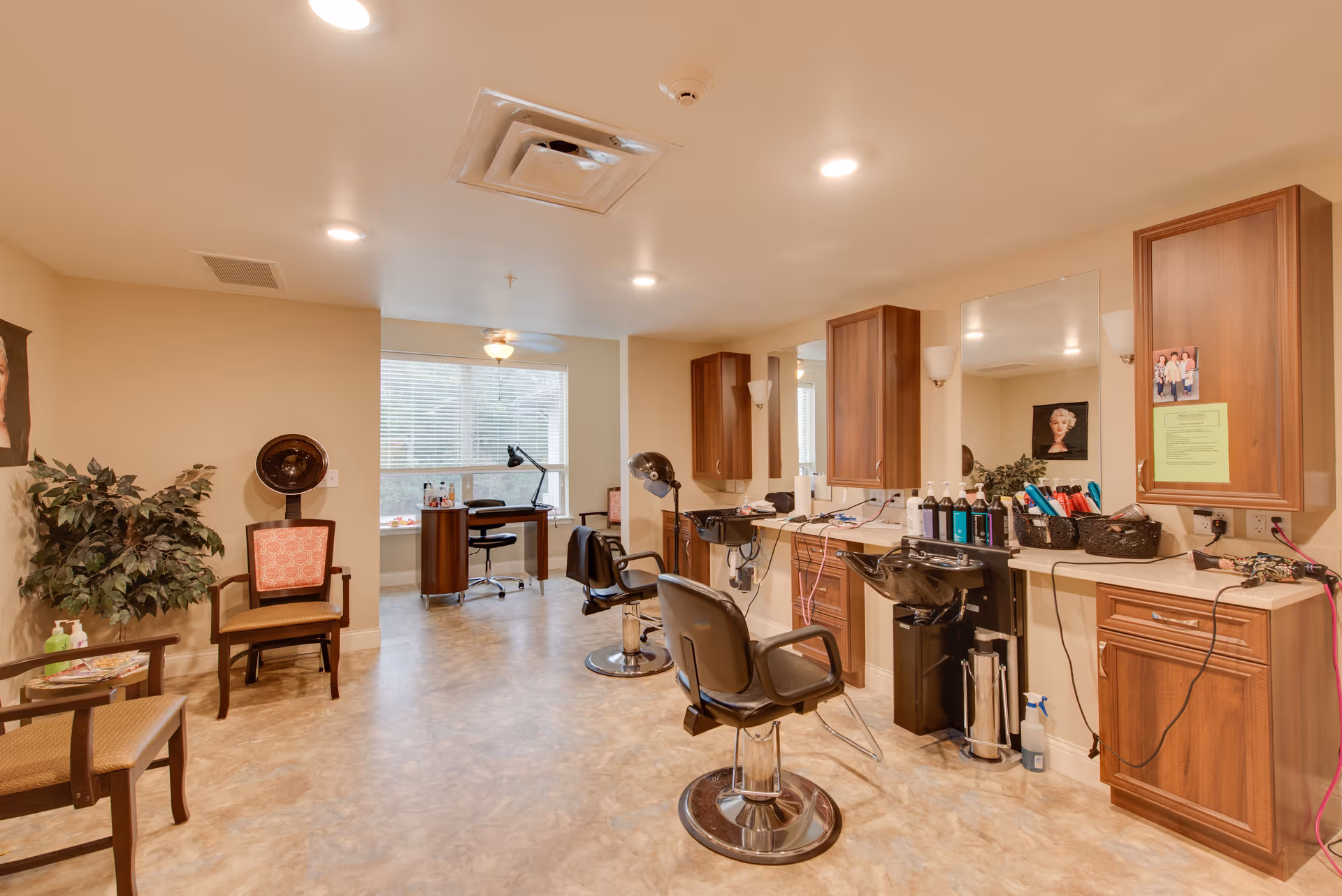 Interior view of a hair salon area in a senior living facility with two salon chairs in front of mirrors and sinks, wooden cabinets, hair care products on the counter, a hair dryer chair, and a desk with a chair near a window in the background.
