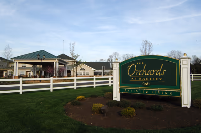 Exterior view of The Orchards Assisted Living facility showing a large green and gold sign with the facility name in front of a white fence and landscaped lawn, with the building and a covered entrance visible in the background under a blue sky.