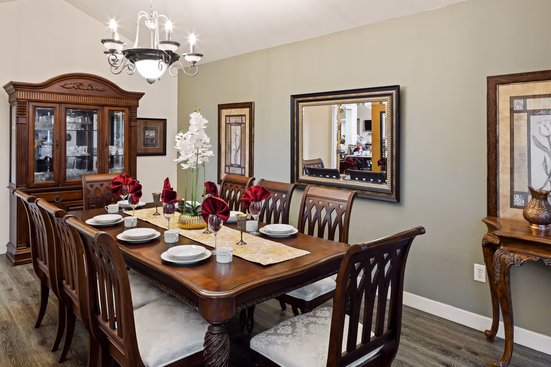 A formal dining room with a large wooden dining table set for eight people. The table features white plates, cups, wine glasses, and red folded napkins. A decorative centerpiece with white flowers is placed in the middle. The room has a wooden china cabinet filled with glassware, a chandelier overhead, a large mirror on the wall, and framed artwork. The floor is wood, and the walls are painted light green.