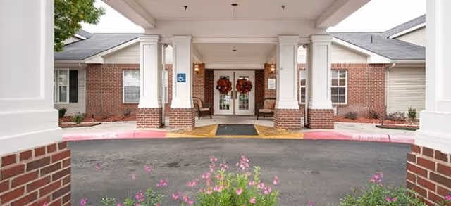 Entrance of a senior living facility with a covered drop-off area supported by white columns with brick bases. The building has beige siding and brick accents, with double glass doors decorated with red wreaths. There are two chairs on either side of the entrance and a handicapped parking sign visible on one of the columns. Pink flowers are in the foreground near the driveway.