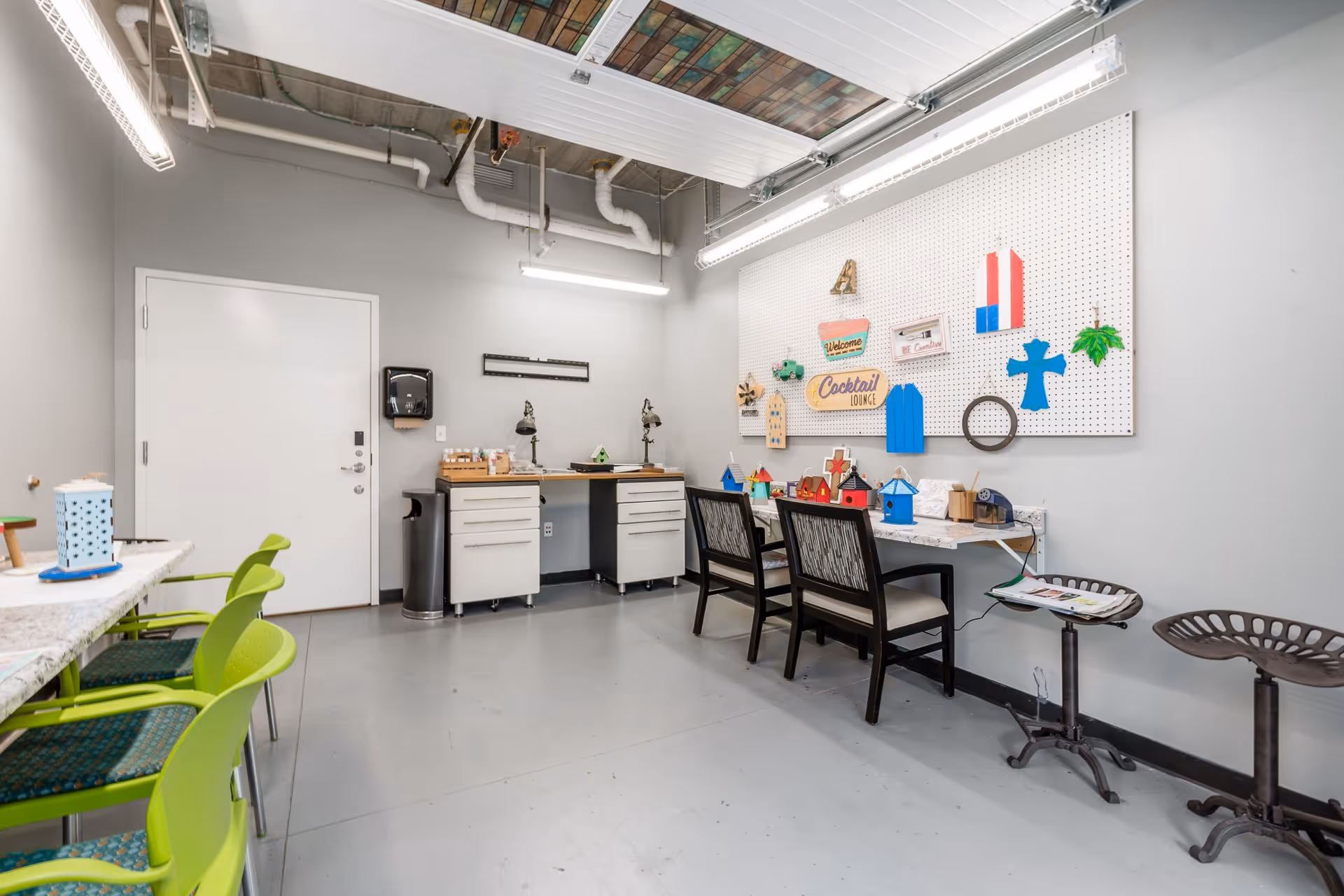 A craft room with gray walls and floor, featuring a pegboard on the wall with various colorful wooden decorations and signs. There are two black chairs with patterned cushions at a counter workspace, two industrial-style stools, and a table with green chairs on the left side. The ceiling has exposed pipes and fluorescent lighting.