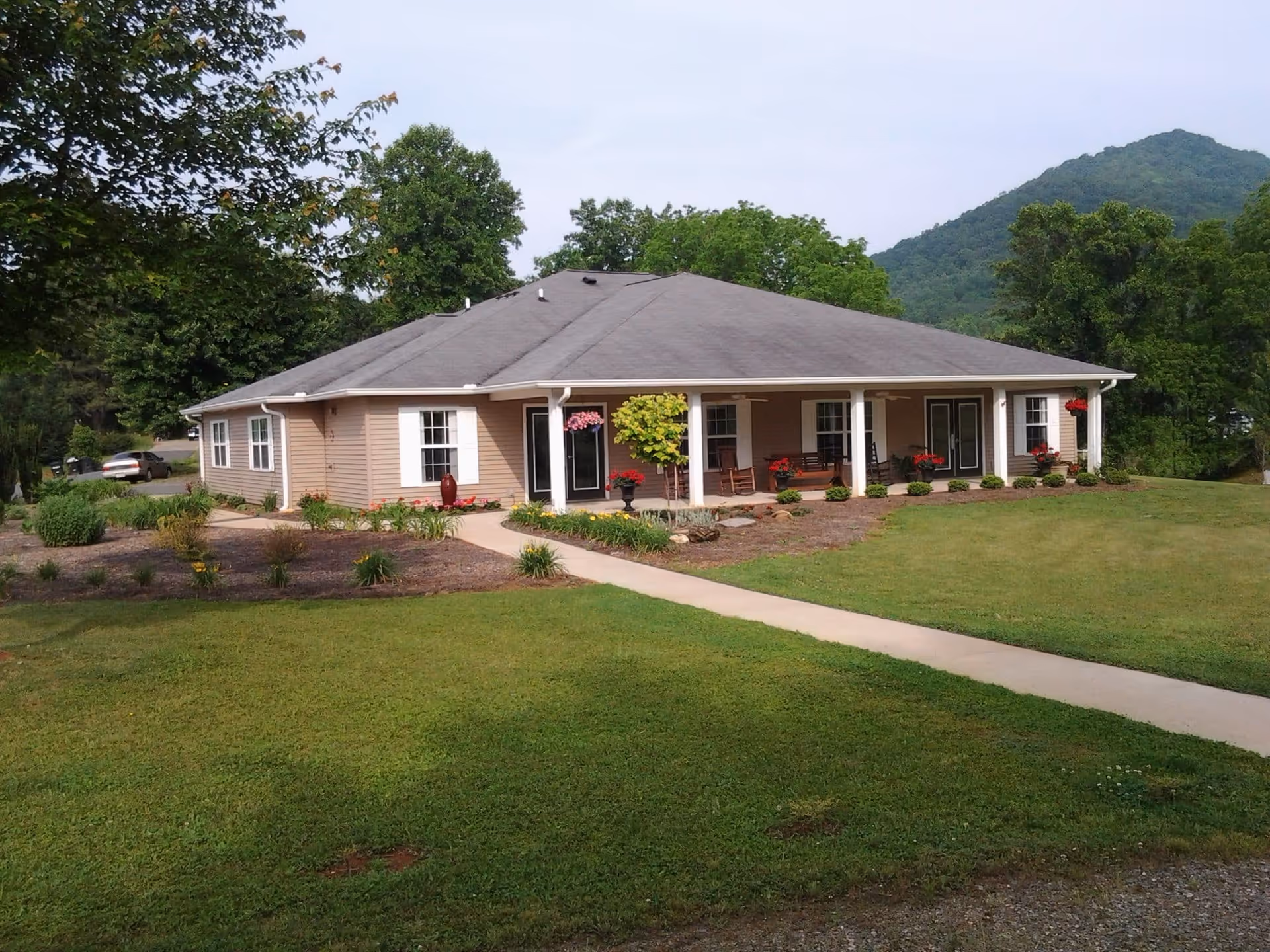Single-story beige building with a gray roof surrounded by green grass and trees, featuring a covered porch with rocking chairs and flower pots, set against a backdrop of a forested hill.