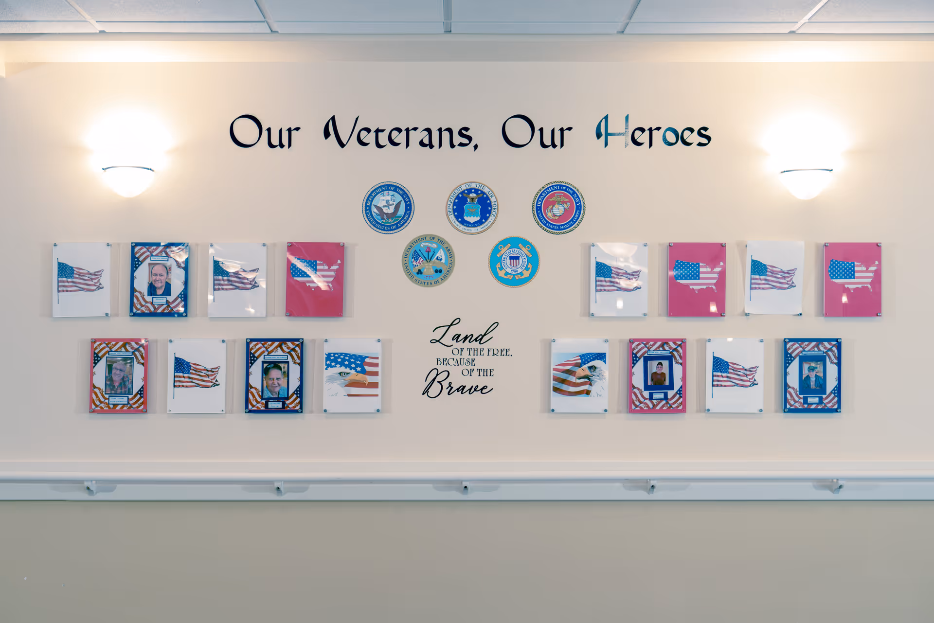 A wall display honoring veterans with the text 'Our Veterans, Our Heroes' above several framed photos and American flag images. Below the photos are military branch emblems and the phrase 'Land of the Free, Because of the Brave'. The wall is softly lit by two wall sconces.
