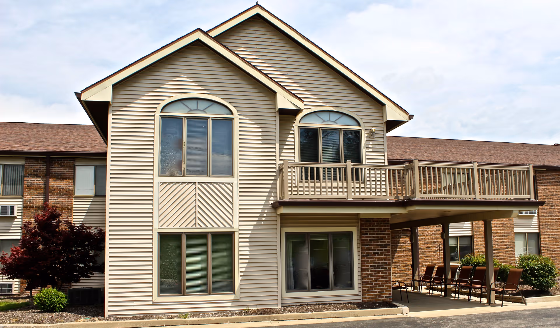 Two-story beige and brick senior living building with a second-floor balcony, arched windows, and outdoor seating under a covered patio.