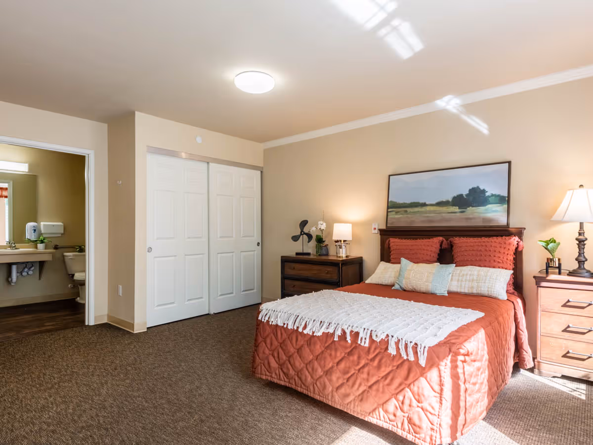 Sunlit bedroom with a made bed, nightstands and dresser, closet doors, and an open doorway to a bathroom.