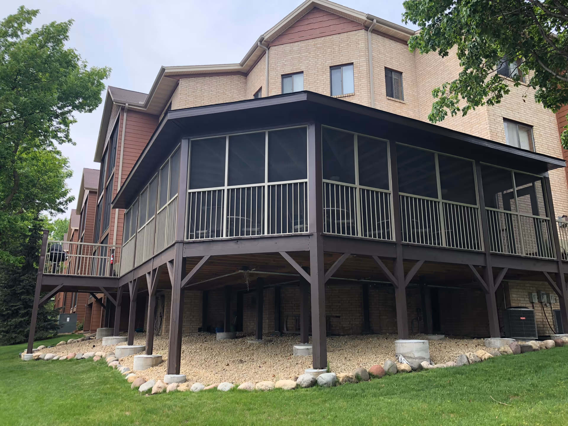 Exterior view of a multi-story senior living facility building with a screened-in porch elevated on wooden supports. The building has beige brick and brown siding, surrounded by green grass and trees under a cloudy sky.
