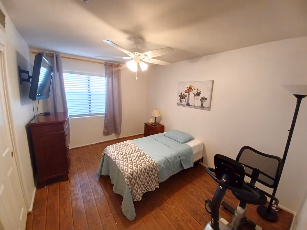 Small bedroom with a single bed, wooden floors, a ceiling fan, wall-mounted TV, dresser, nightstand, and an exercise bike.