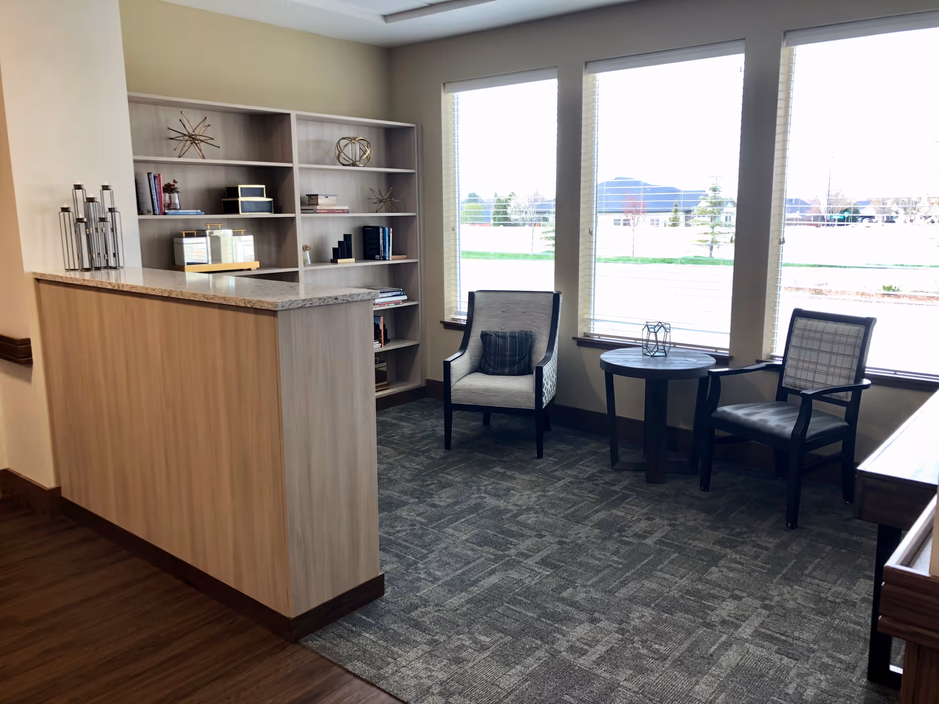 Sunlit seating area with two armchairs, a small round table, bookshelf, and a counter by large windows overlooking the outdoors.
