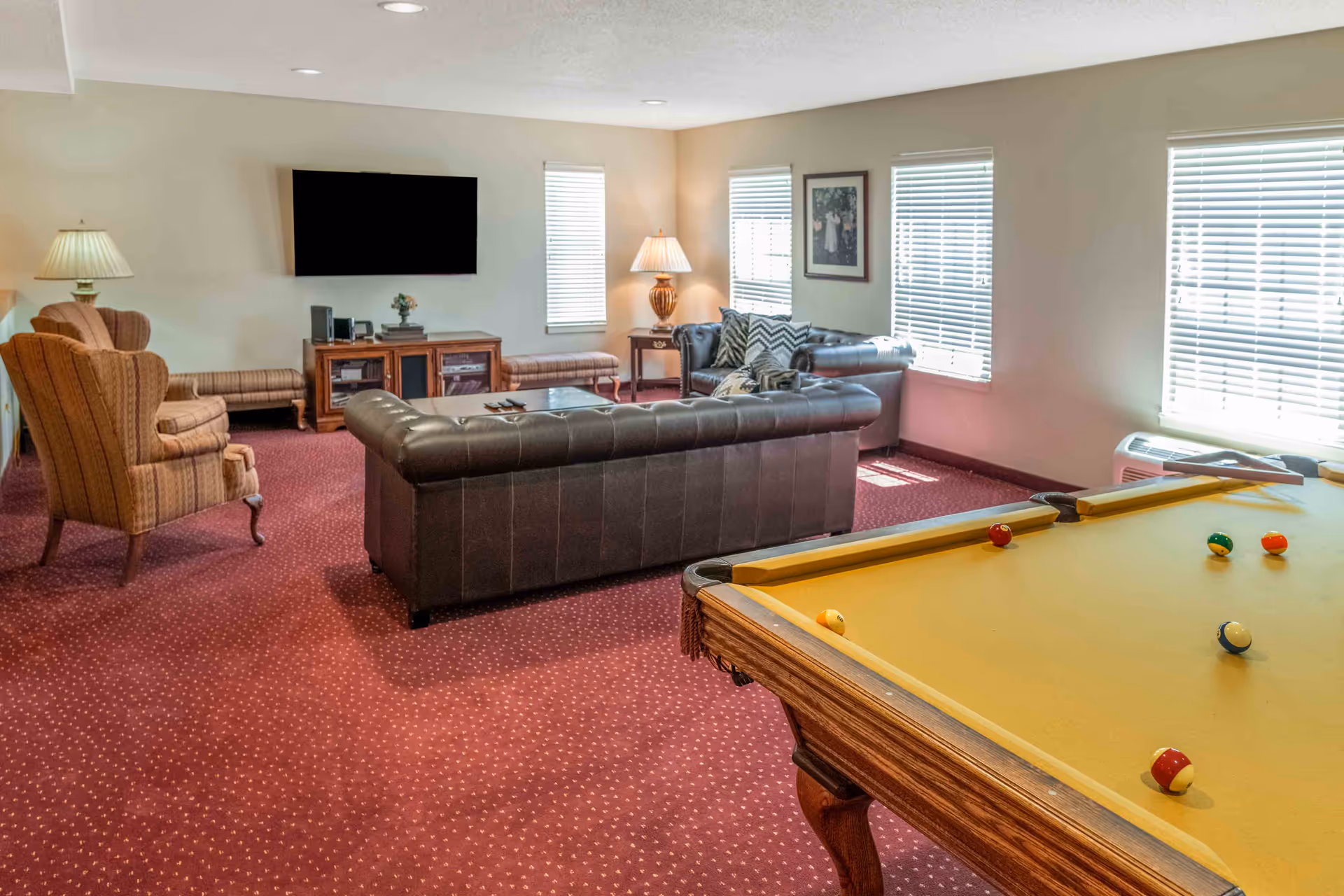 A carpeted common room with leather sofas, armchairs, a wall-mounted TV, lamps, and a pool table in the foreground.