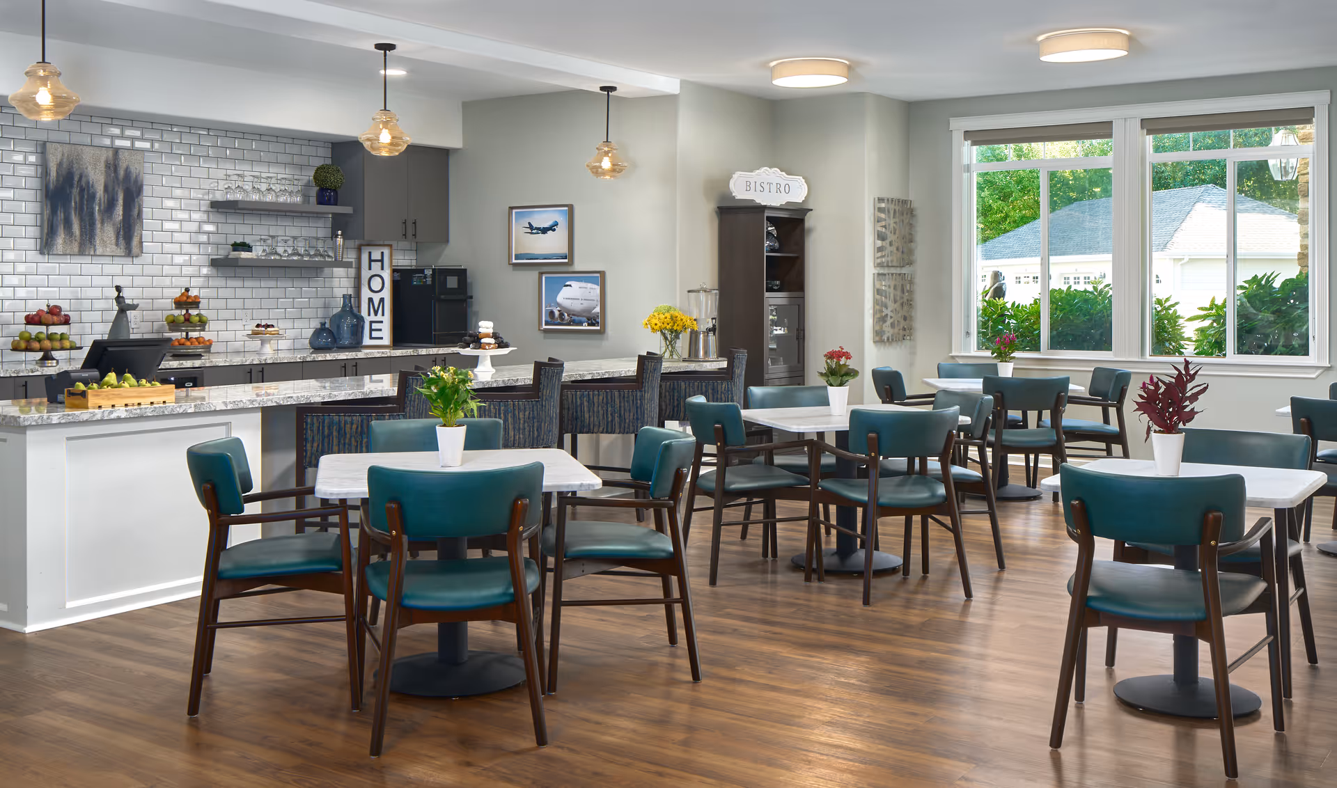 A bright and modern dining area with multiple tables and teal chairs, each table decorated with small potted plants. The room features large windows letting in natural light, a kitchen counter with granite countertops, pendant lights hanging from the ceiling, and a cabinet labeled 'Bistro'. The kitchen area has white subway tile backsplash, shelves with glassware, and a sign that reads 'HOME'.