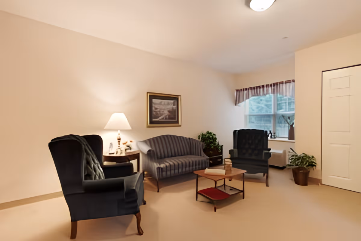 A cozy living room with two dark upholstered armchairs, a striped sofa, a wooden coffee table, a side table with a lamp, a framed picture on the wall, a window with a valance, and two potted plants.