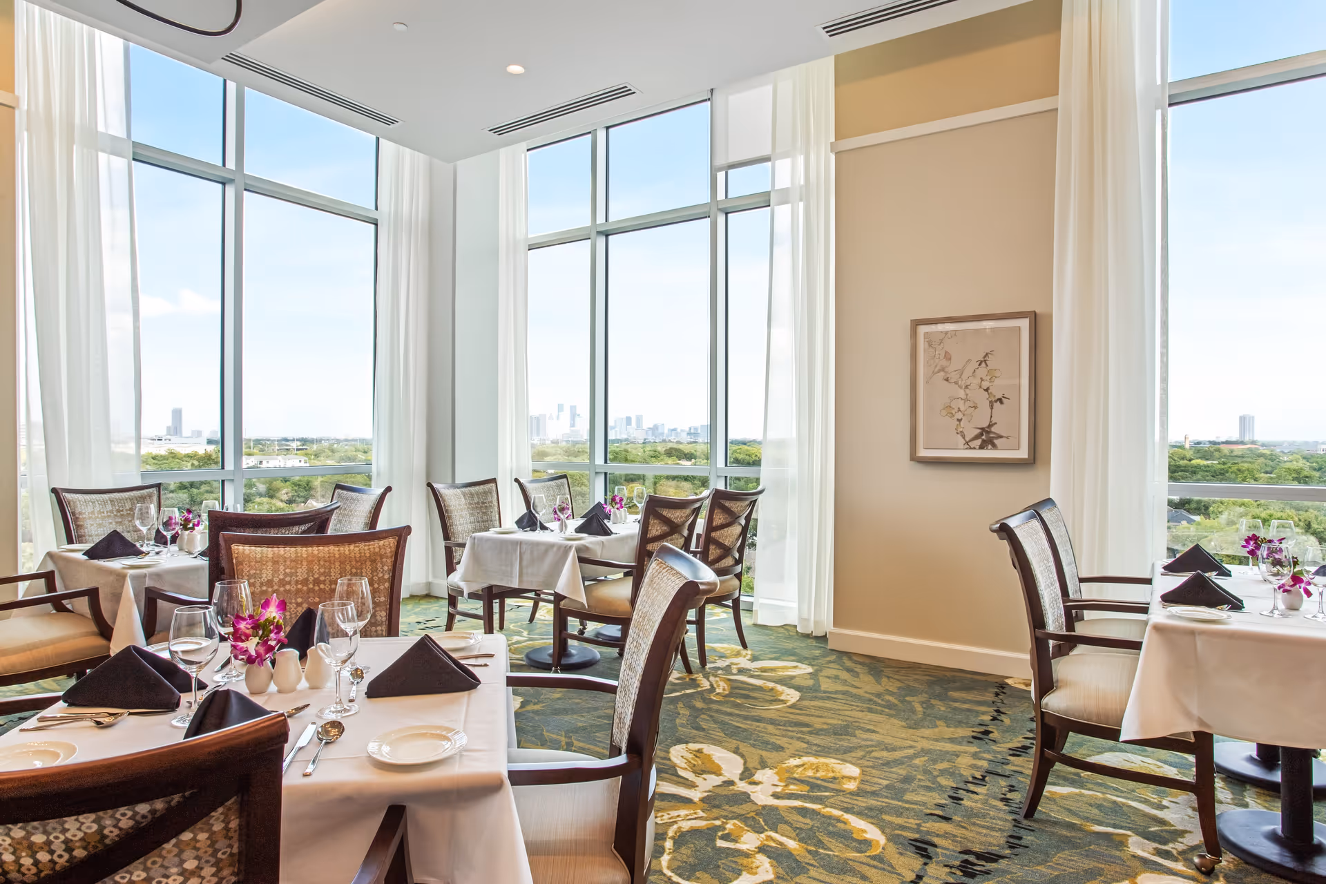 Bright dining room with large floor-to-ceiling windows offering a scenic view of a city skyline and greenery. Several tables are set with white tablecloths, black folded napkins, glassware, plates, and small floral centerpieces. The room has patterned carpet and light-colored walls with a framed artwork.