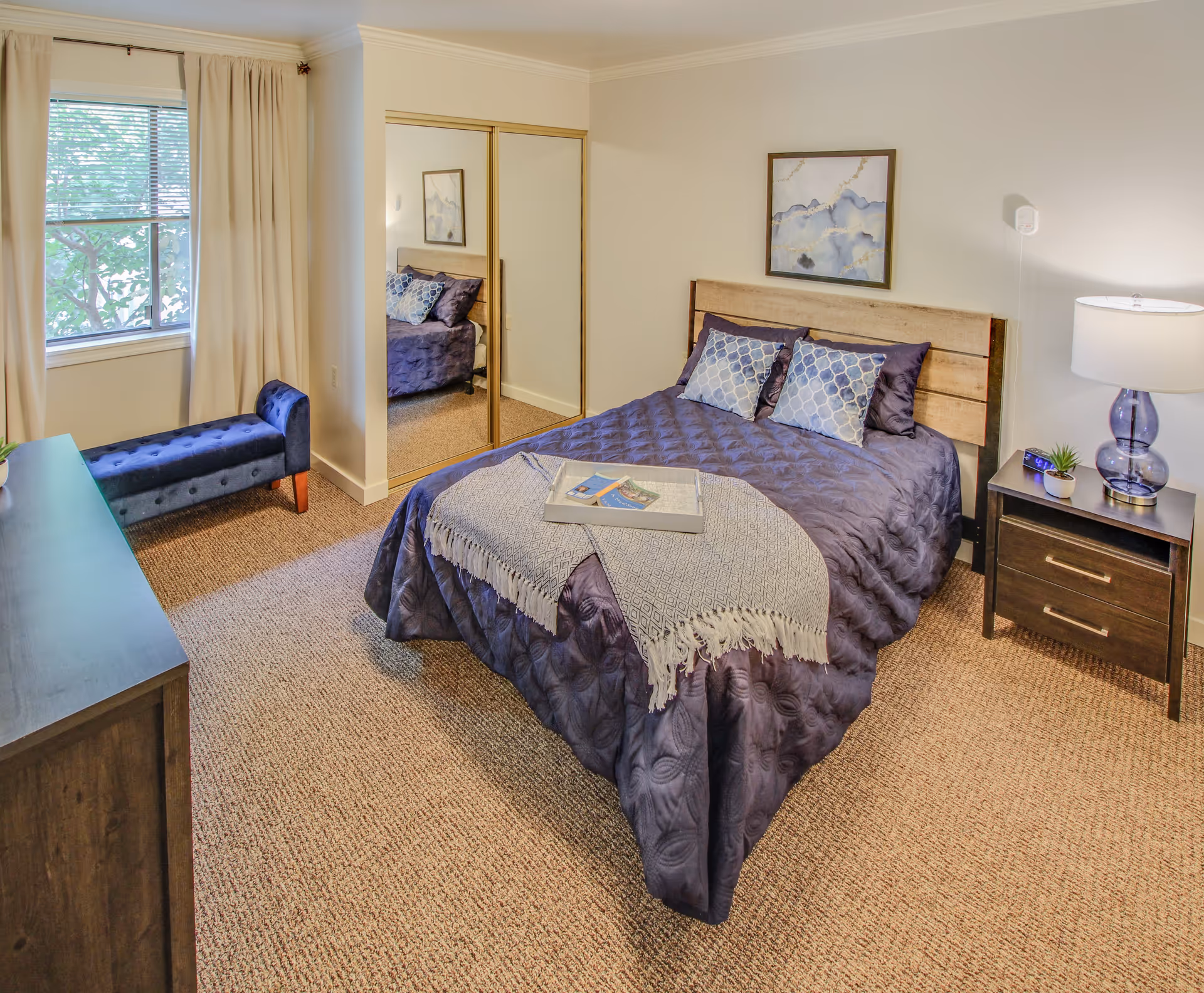A cozy bedroom with a bed covered in a dark purple quilt and two decorative pillows. A light gray throw blanket is draped over the foot of the bed with a tray on top. There is a nightstand with a lamp and small plant on the right side of the bed. A dresser is on the left side of the room, and a blue upholstered bench is placed near a window with beige curtains. A closet with mirrored sliding doors is visible on the far wall, and a framed abstract painting hangs above the bed.