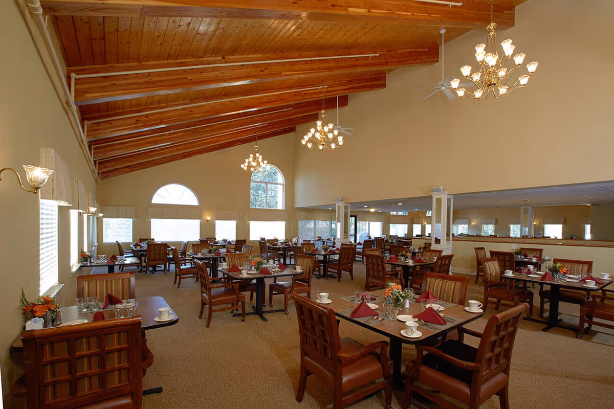 Spacious dining room with a wooden-beamed vaulted ceiling, chandeliers, and multiple tables set with place settings and chairs.
