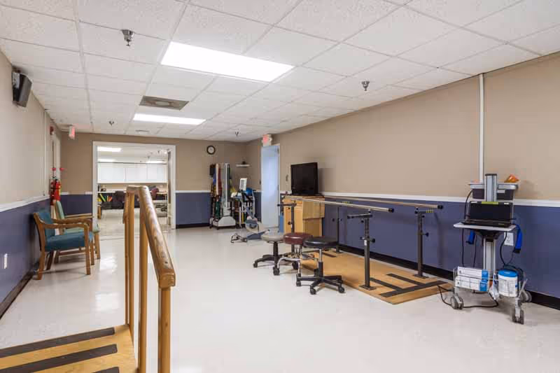 A rehabilitation or physical therapy room with parallel bars for walking exercises, stools, a TV mounted on a stand, and various medical and therapy equipment along the walls. The room has beige and blue walls, a white ceiling with fluorescent lights, and a white floor. There are also chairs along the left wall and an open doorway leading to another room.