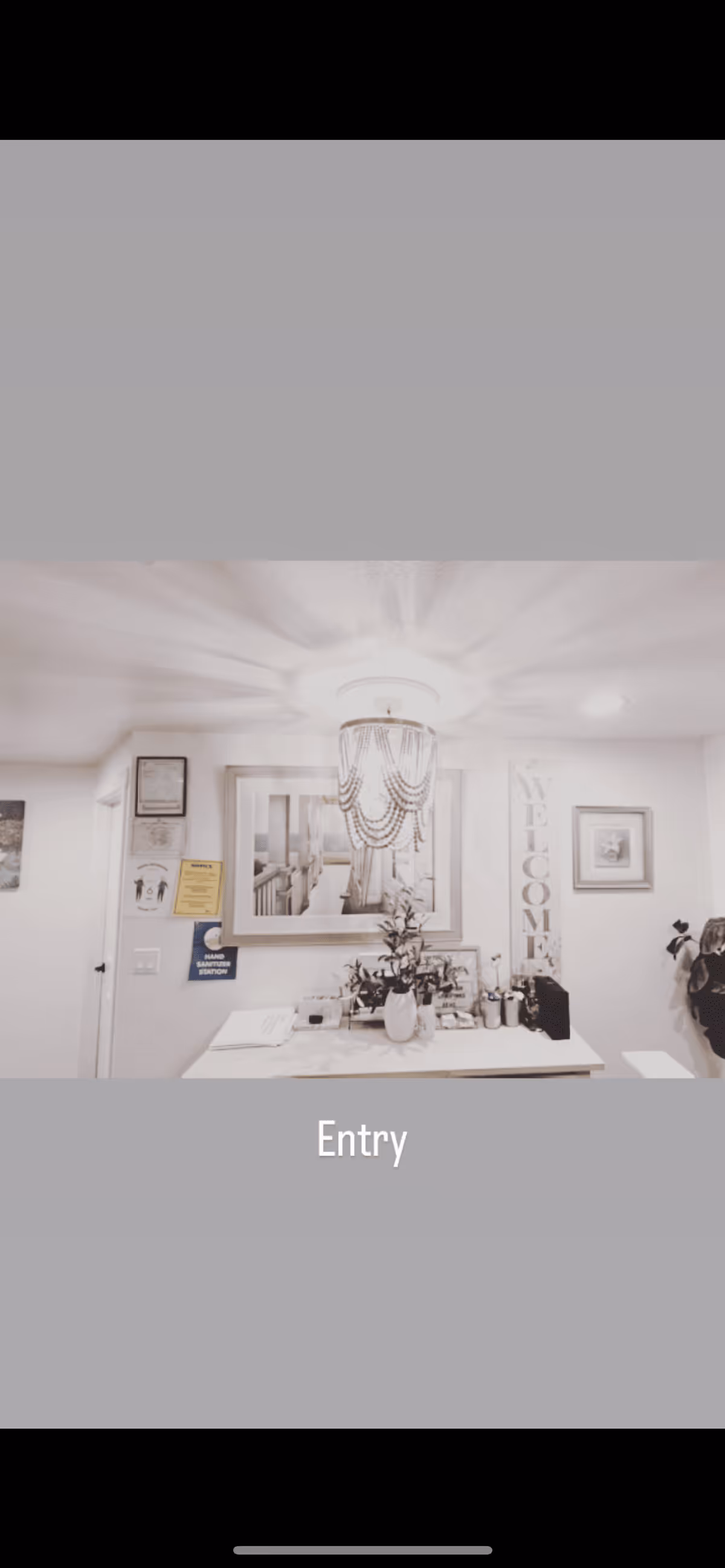 Entry area of Alderwood Elderly Home Care Adult Family Home with a white table holding a vase with flowers and various items. Above the table is a framed picture and a decorative chandelier. On the wall to the right is a vertical 'WELCOME' sign and a framed picture. On the left wall, there are some notices and a hand sanitizing station sign.