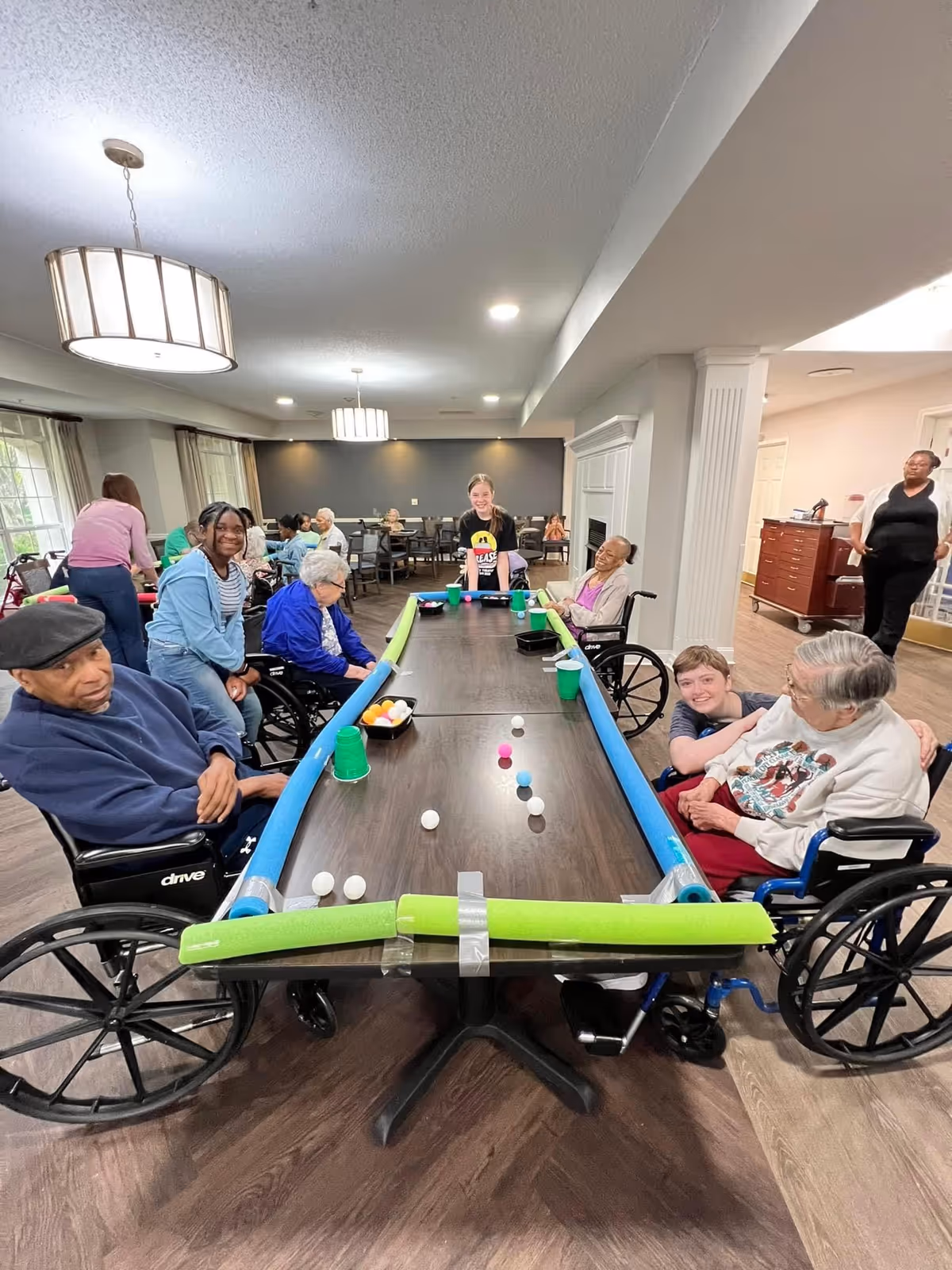 A group of elderly individuals, some in wheelchairs, and a few younger people are gathered around a long table playing a game with ping pong balls and cups in a well-lit common area with modern lighting and wood flooring.